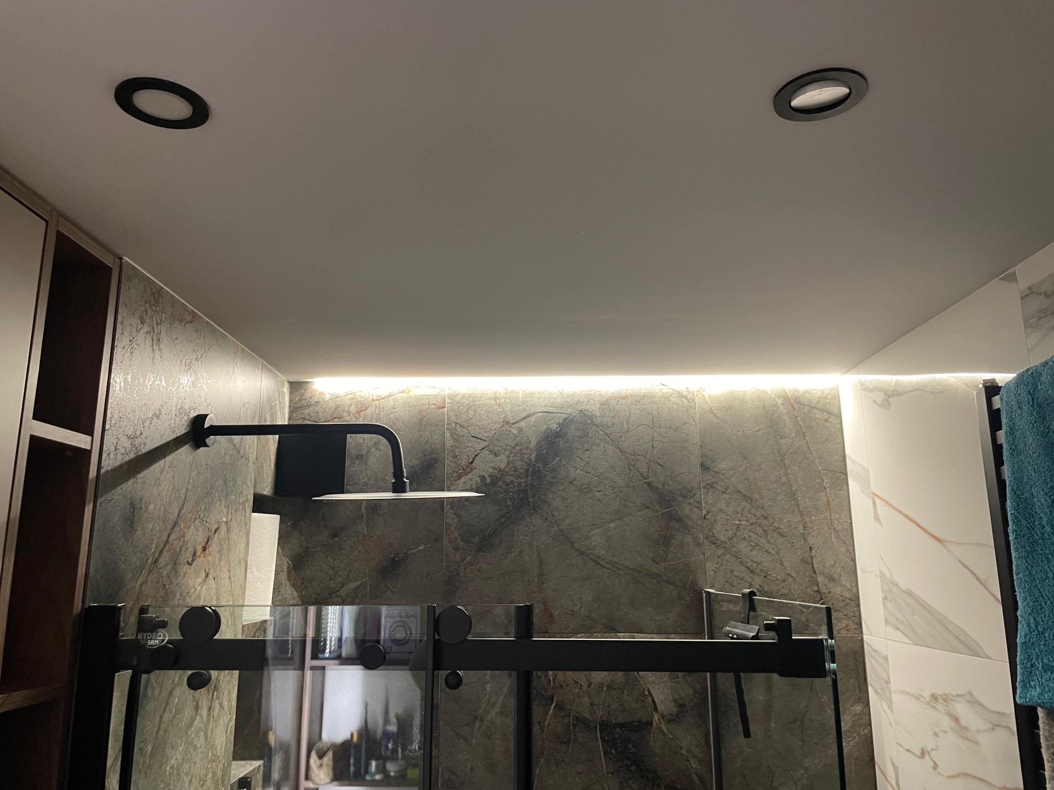 Nowoczesna kabina prysznicowa z czarną armaturą i podświetleniem LED, widoczne dwa okrągłe reflektory w suficie. Marmurowe płytki na ścianach.