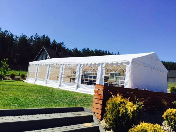 Biały namiot imprezowy z przezroczystymi ścianami na zielonym trawniku, widok z lekkiej góry, schody w kadrze, drzewa w tle, błękitne niebo.