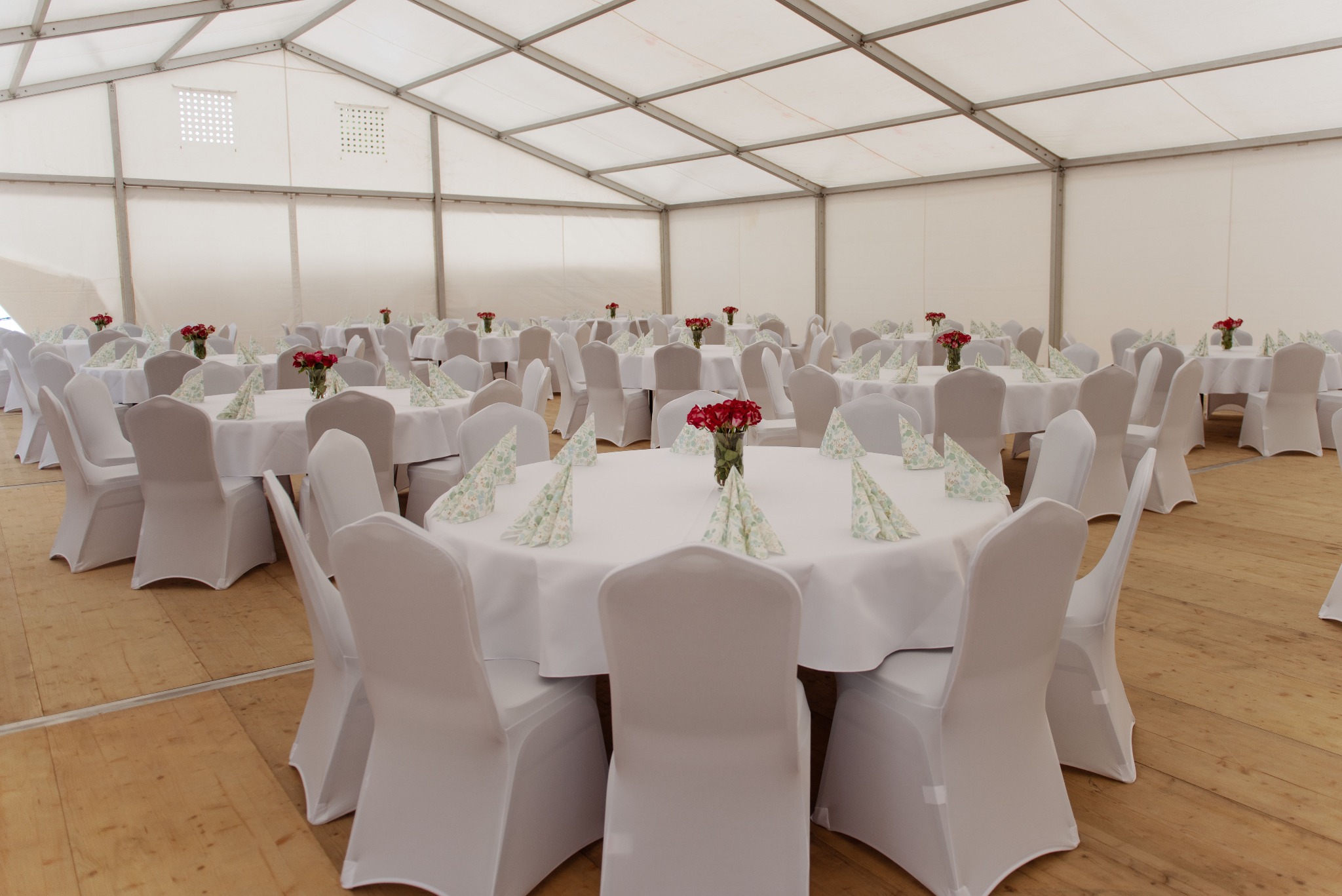 Uroczyste wnętrze białego namiotu bankietowego z okrągłymi stołami, krzesłami w pokrowcach i dekoracjami z czerwonych róż. Drewniana podłoga, jasne oświetlenie.