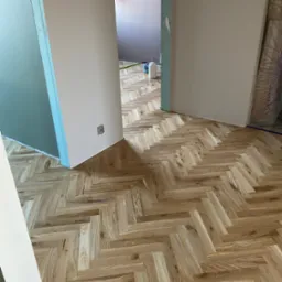 Ułożony parkiet w jodełkę francuską w jasnym odcieniu drewna, widok z perspektywy podłogi, ściany w trakcie malowania, gniazdko elektryczne na ścianie.