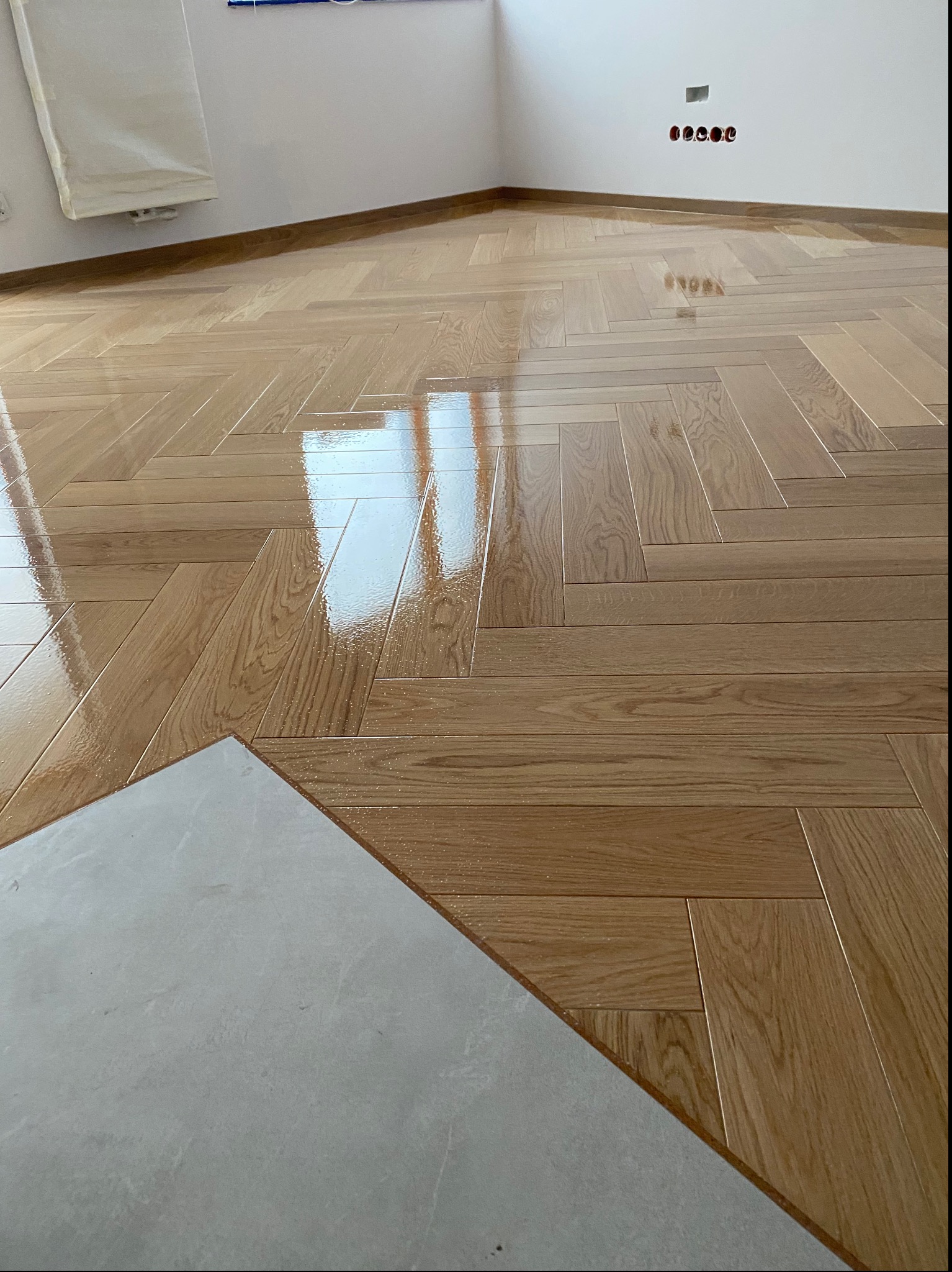 Świeżo polakierowany parkiet ułożony w jodełkę, z widocznym fragmentem szarej płytki podłogowej w rogu pomieszczenia.
