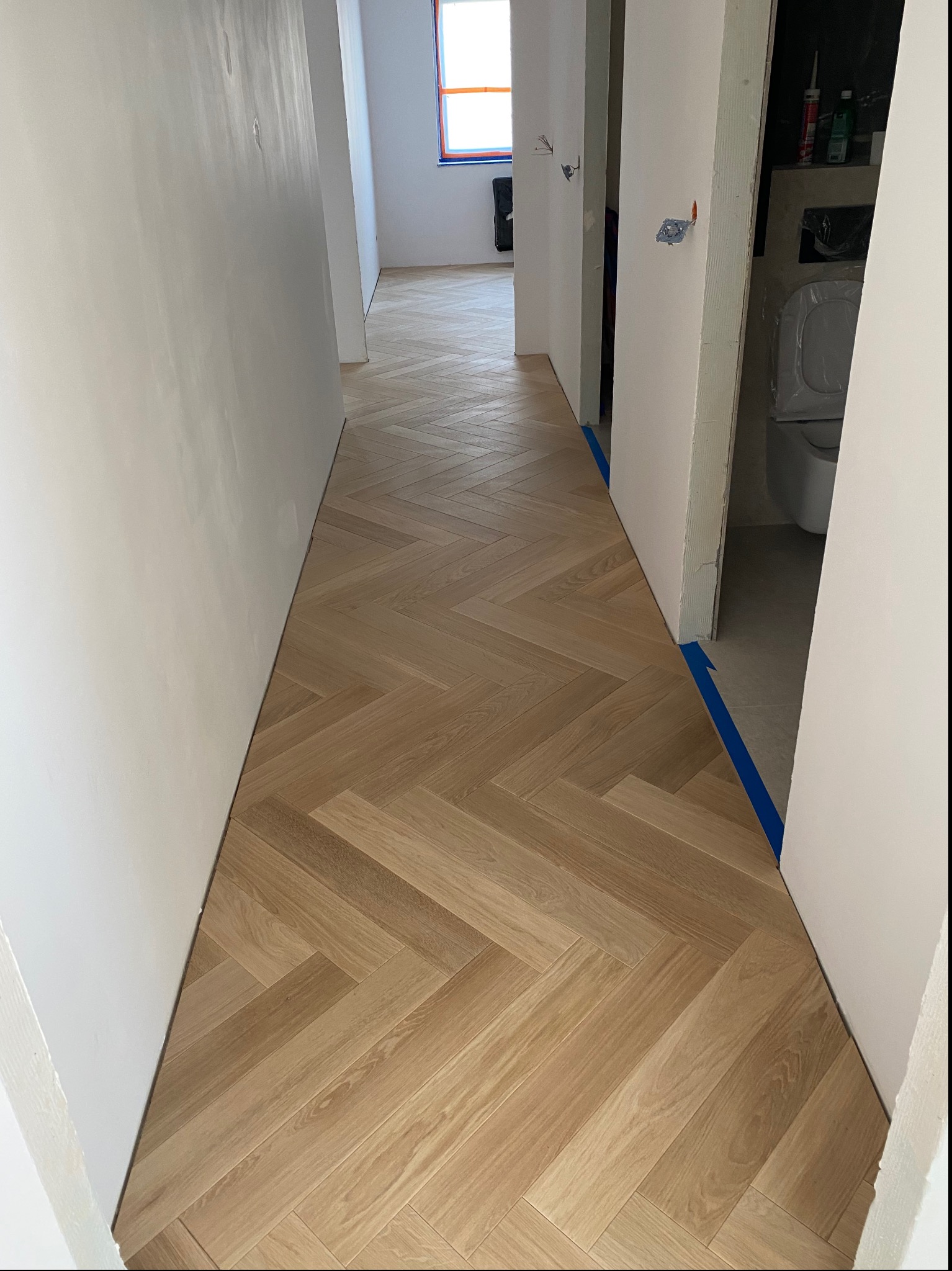 Nowo ułożony parkiet w jodełkę w korytarzu, zabezpieczony niebieską taśmą malarską przy wejściu do łazienki, ściany przygotowane do malowania.
