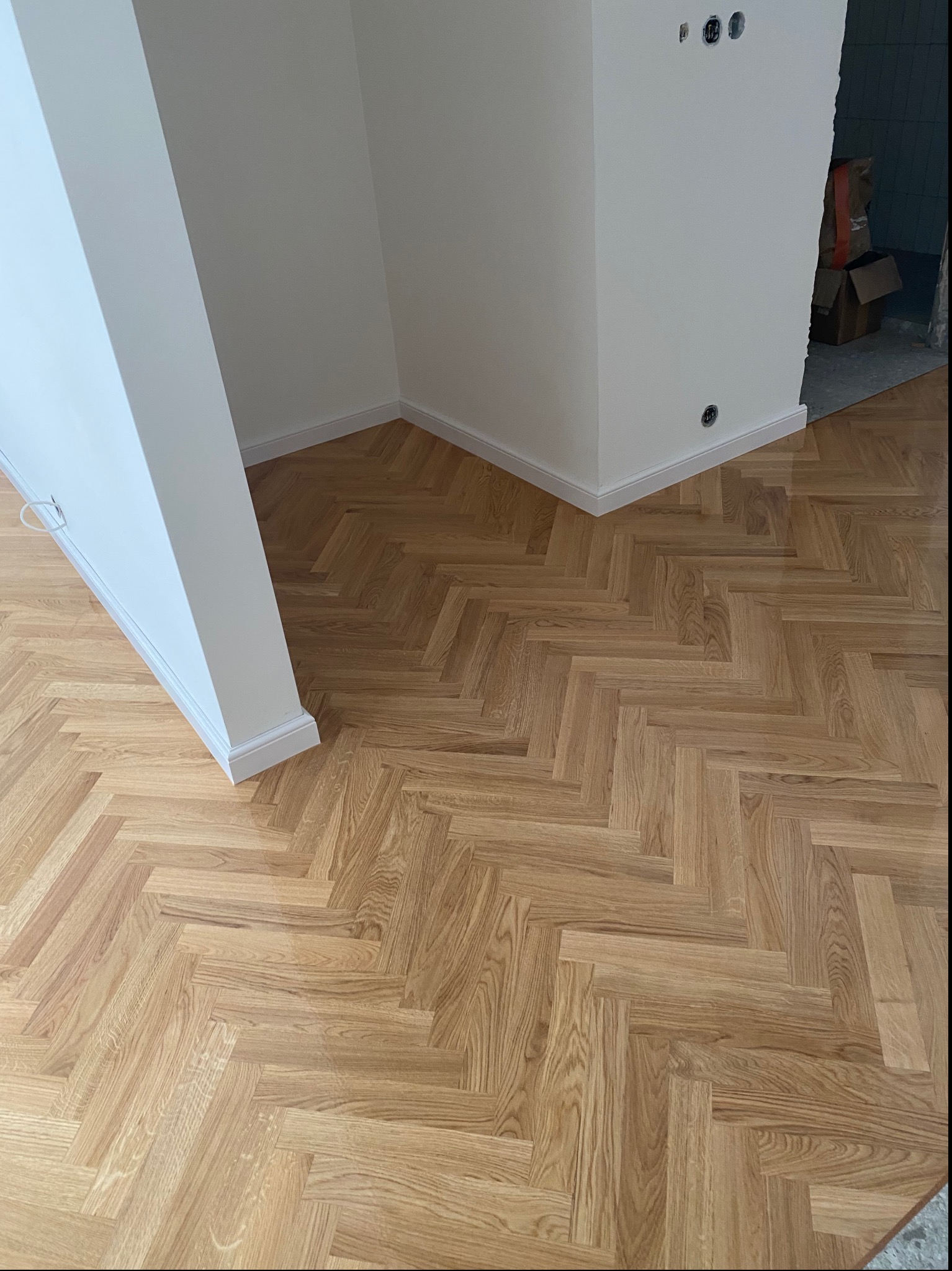 Ułożony parkiet w jodełkę klasyczną, jasne drewno, białe listwy przypodłogowe, widoczne puszki elektryczne w ścianie, stan surowy w przejściu do innego pomieszczenia.