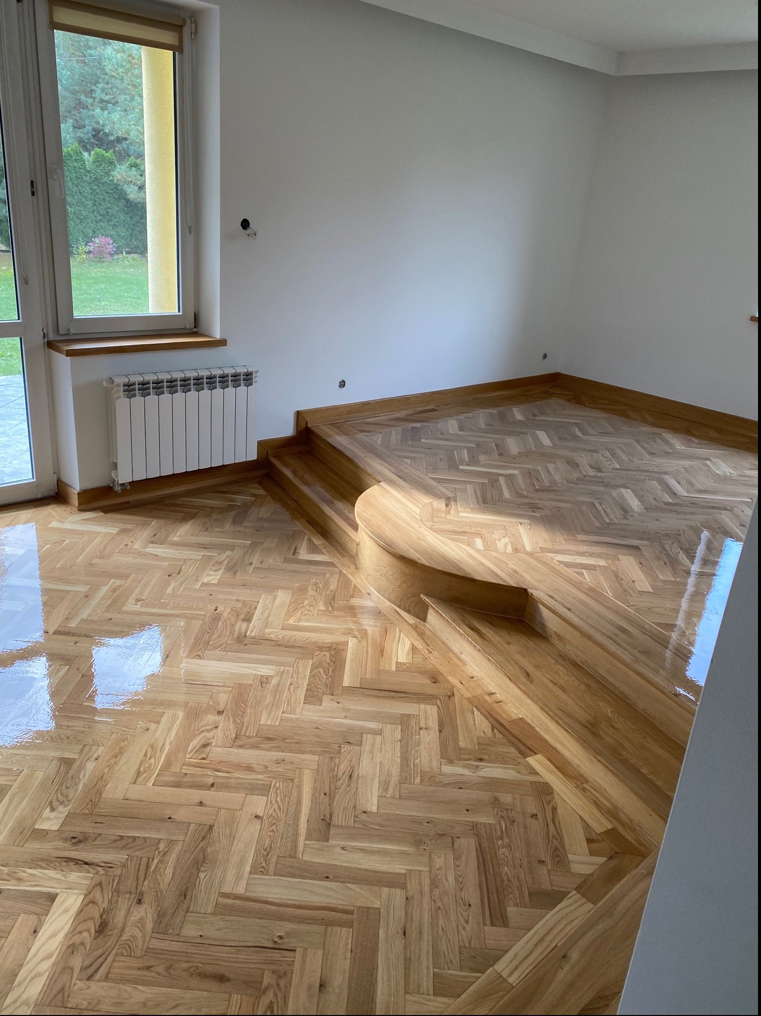 Świeżo polakierowany parkiet ułożony w jodełkę na podłodze i schodach, widoczne refleksy światła, białe ściany i grzejnik pod oknem.