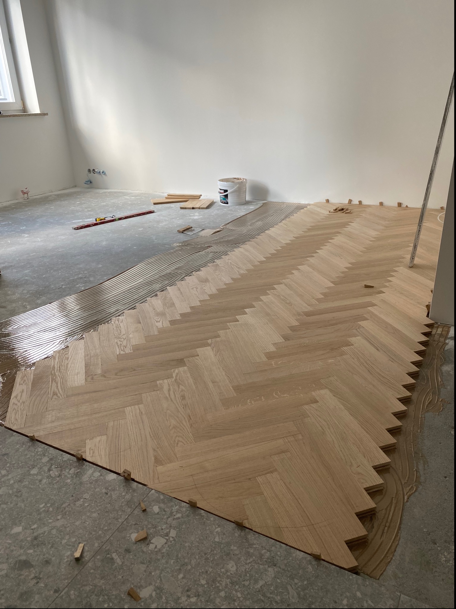 Układanie parkietu w jodełkę francuską, widoczne świeżo nałożone pasy kleju, drewniane kliny dystansowe i poziomica w tle.