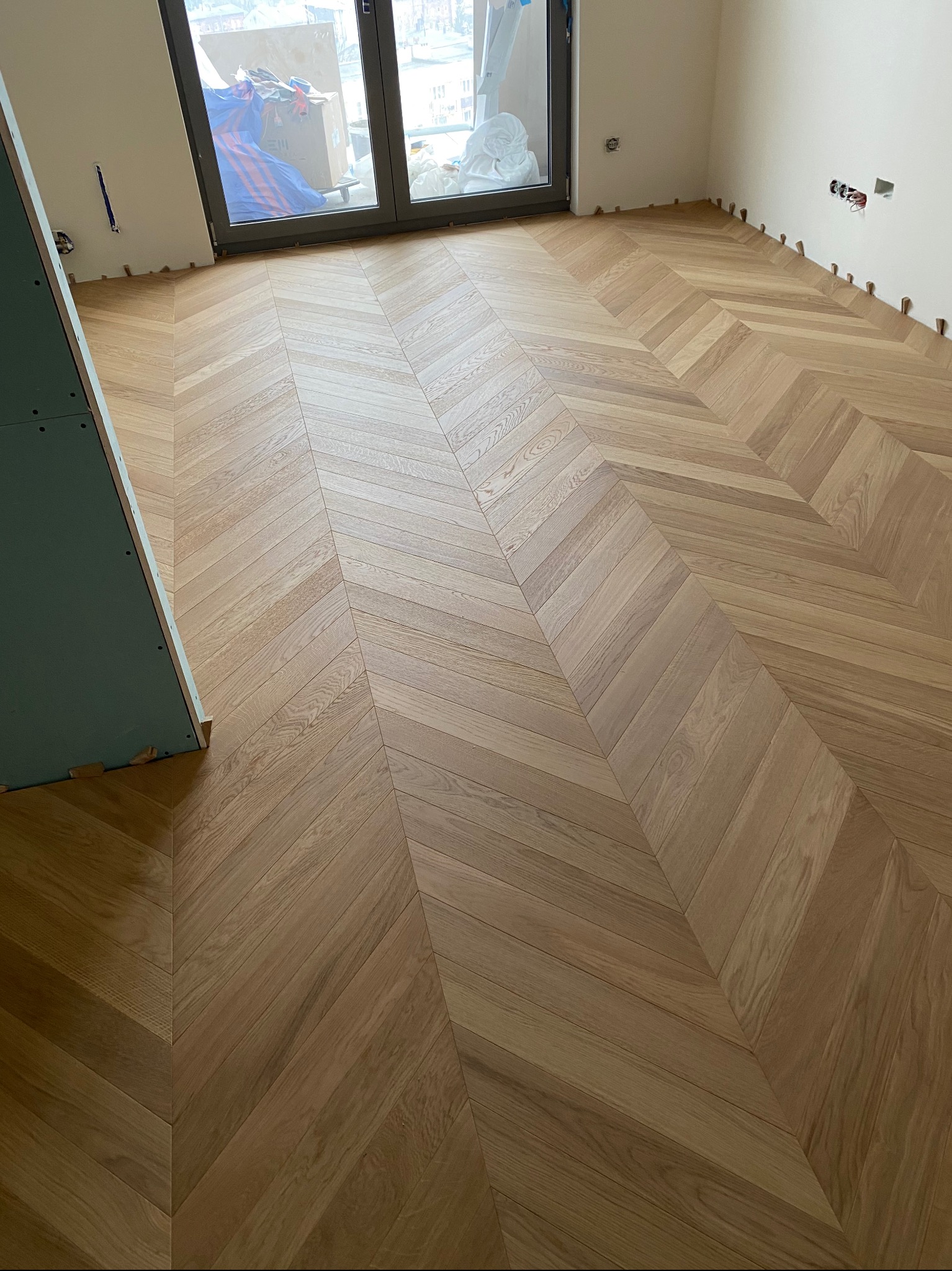 Ułożony parkiet w jodełkę francuską, widoczne listwy dystansowe przy ścianach i fragment płyty gipsowej, okno w tle.