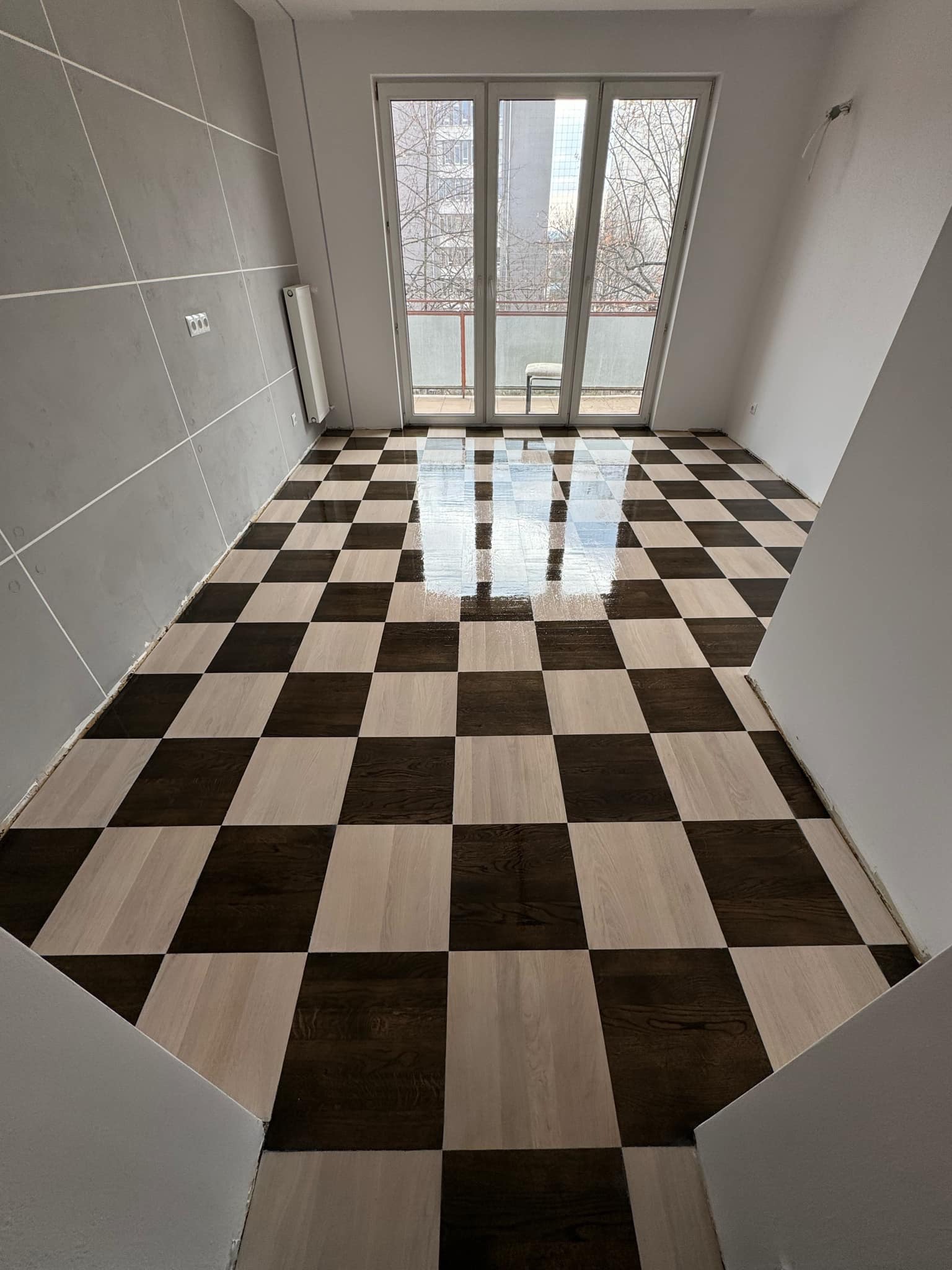 Nowoczesne wnętrze z błyszczącą, szachownicową podłogą z parkietu w odcieniach brązu i beżu, odbijającą światło z okna balkonowego. Szare ściany z białymi listwami.