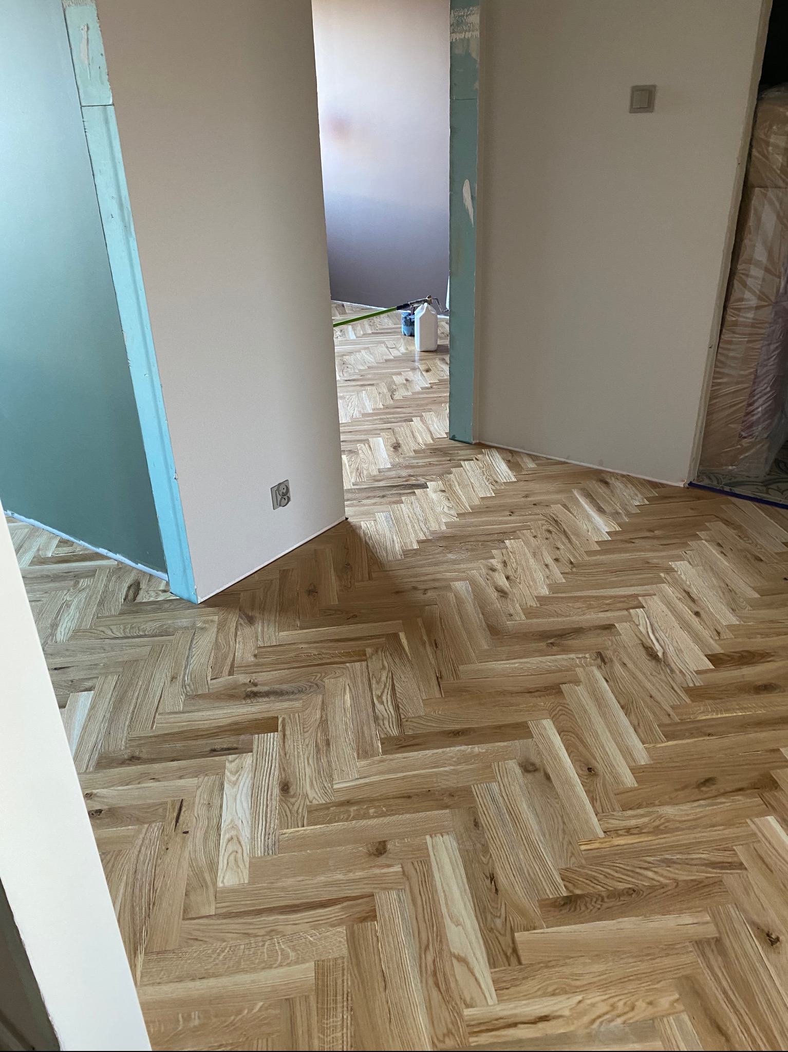 Ułożony parkiet w jodełkę francuską w jasnym odcieniu drewna, widok z perspektywy podłogi, ściany w trakcie malowania, gniazdko elektryczne na ścianie.