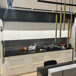 Kmeble usługi stolarskie Krzysztof Nowakowski - Nowoczesna kuchnia z białymi szafkami dolnymi z delikatnymi złotymi uchwytami, czarnymi szafkami górnymi i czarnym blatem, oświetlona złotymi lampami wiszącymi, widoczne naczynia i owoce na blacie.