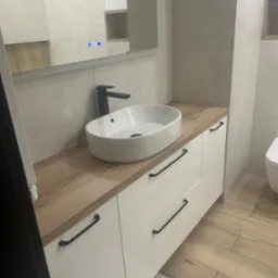 Nowoczesna łazienka: biała, owalna umywalka nablatowa z czarną armaturą, szafka pod umywalkę z białymi, lakierowanymi frontami i drewnianym blatem, drewnopodobne płytki na podłodze, duże lustro...