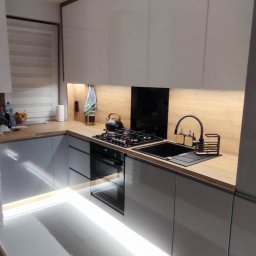 Kmeble usługi stolarskie Krzysztof Nowakowski - Nowoczesna kuchnia w szarościach z drewnianym blatem, oświetleniem LED pod szafkami i na podłodze, czajnik na płycie gazowej.