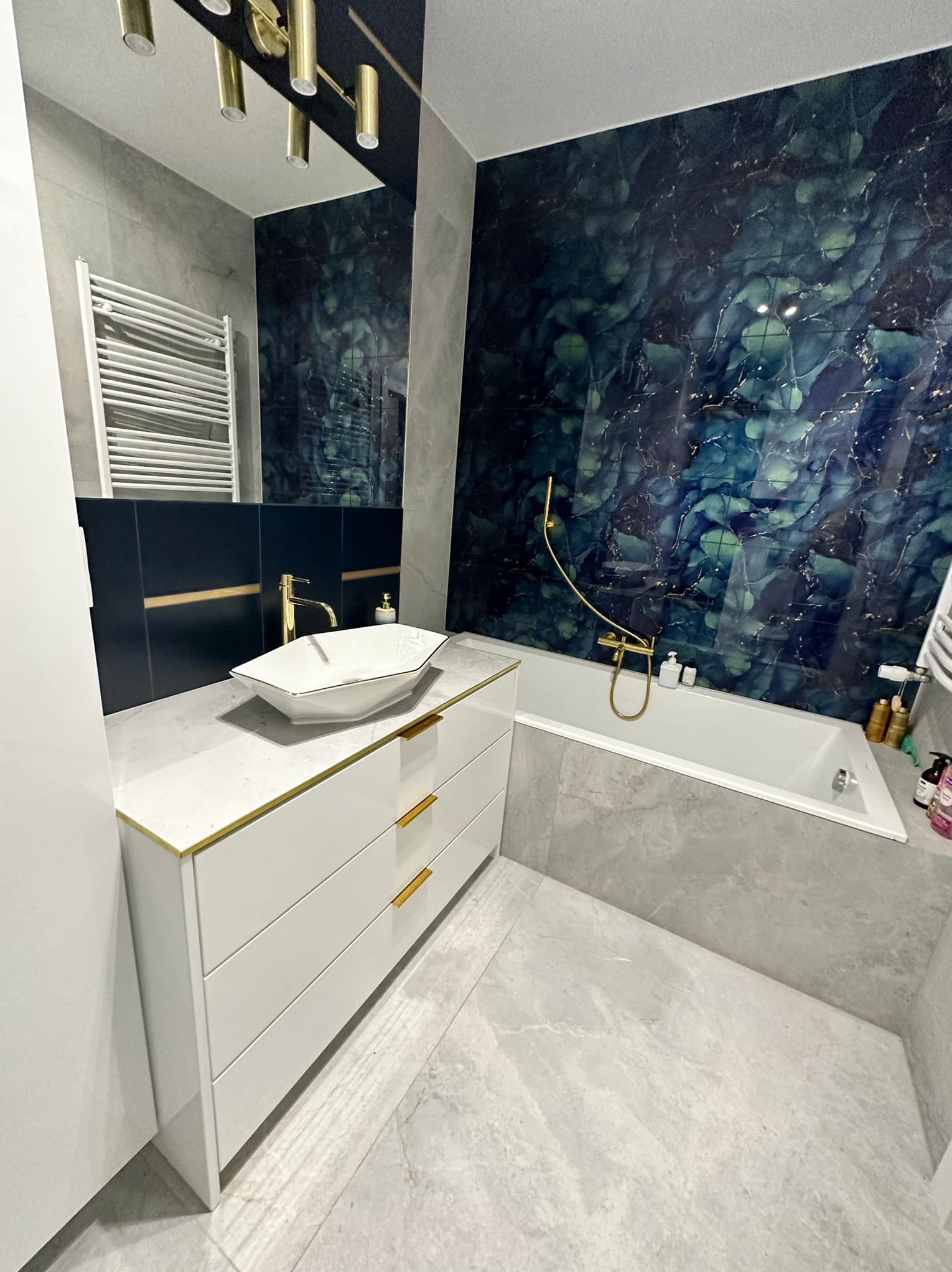 Nowoczesna łazienka z białą szafką pod umywalkę z złotymi akcentami, umywalką w kształcie łódki, baterią w kolorze złotym, wanną i ścianą z efektem marmuru w odcieniach niebieskiego i zieleni.