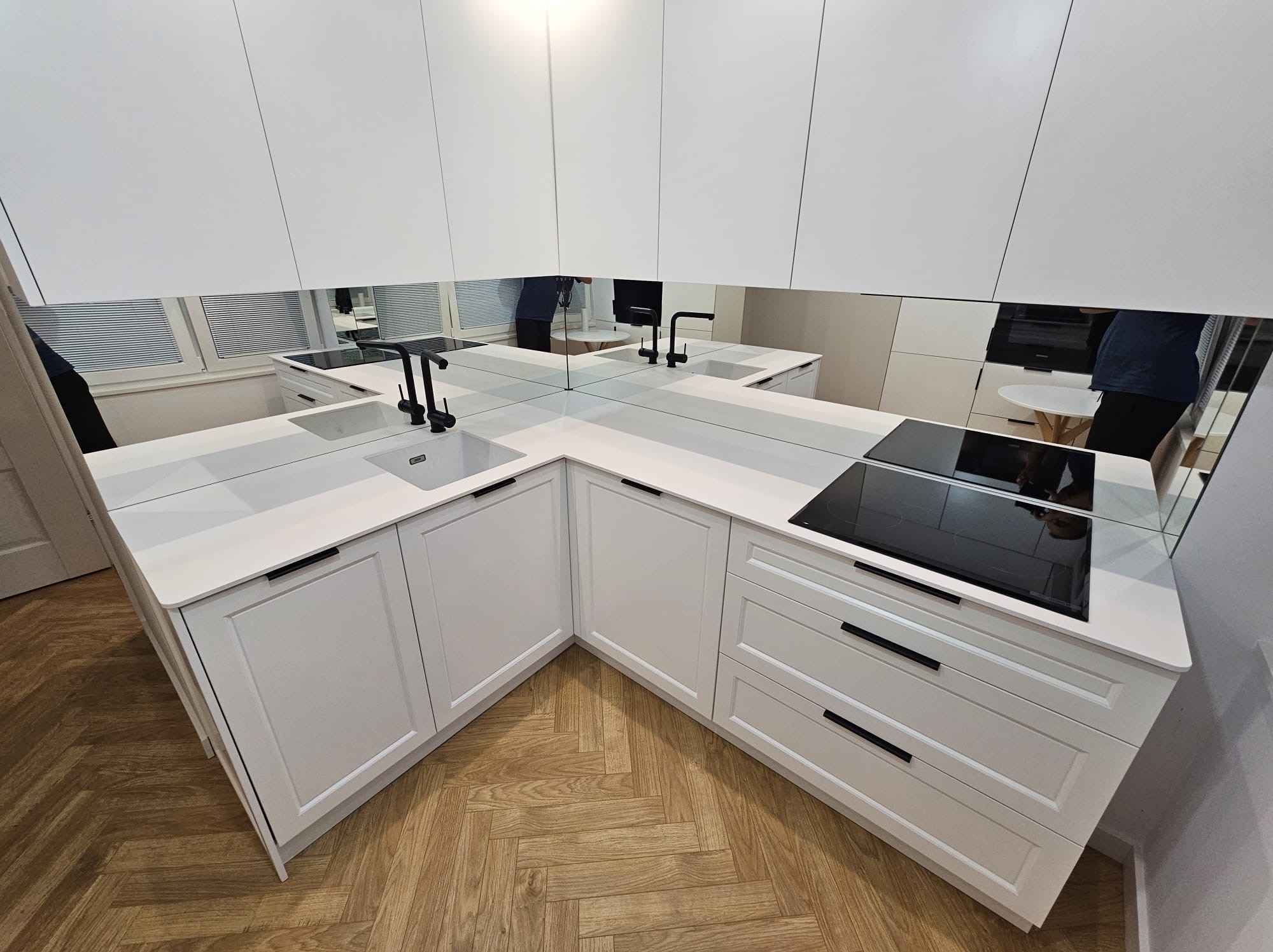 Nowoczesna, biała kuchnia z lustrzanymi panelami nad blatem, czarnymi uchwytami i armaturą, widoczna w Płocku. Podłoga wyłożona parkietem w jodełkę.