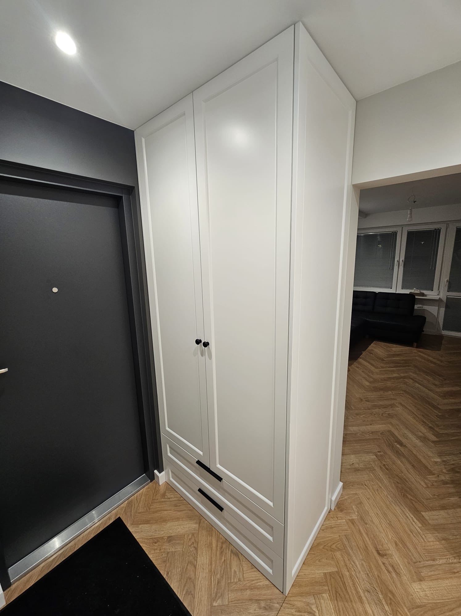 Biała szafa wnękowa z czarnymi uchwytami, umieszczona w przedpokoju z parkietem w jodełkę, widok na salon z czarną sofą.