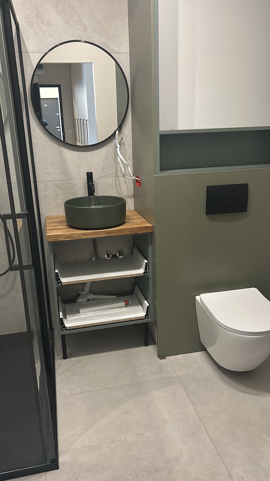 Nowoczesna łazienka z okrągłą umywalką w odcieniach zieleni, drewnianym blatem i otwartą szafką z widocznymi elementami instalacji hydraulicznej oraz wiszącą toaletą z czarnym przyciskiem.