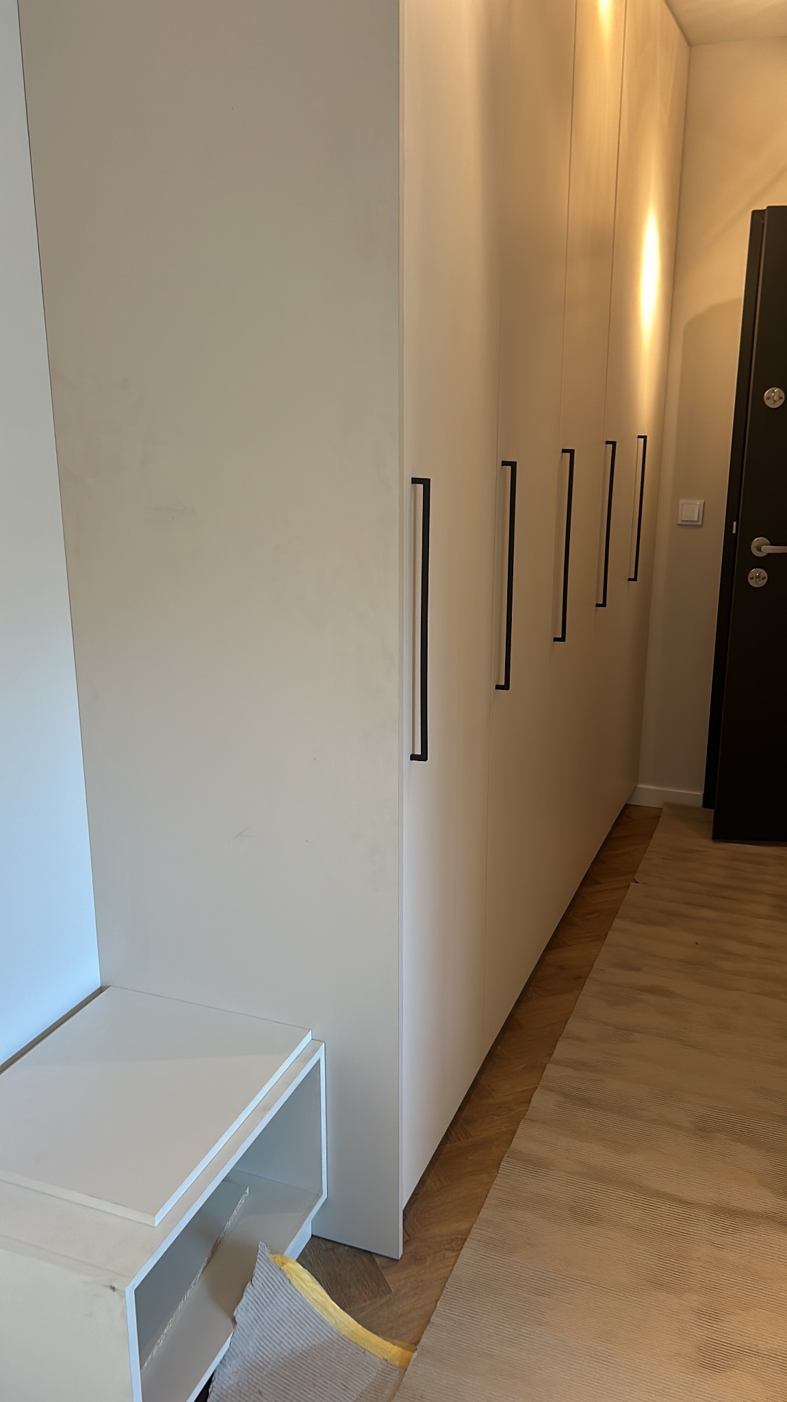 Biała szafa wnękowa z długimi, czarnymi uchwytami, dopasowana do zabudowy przedpokoju z siedziskiem i drewnianą podłogą w jodełkę.