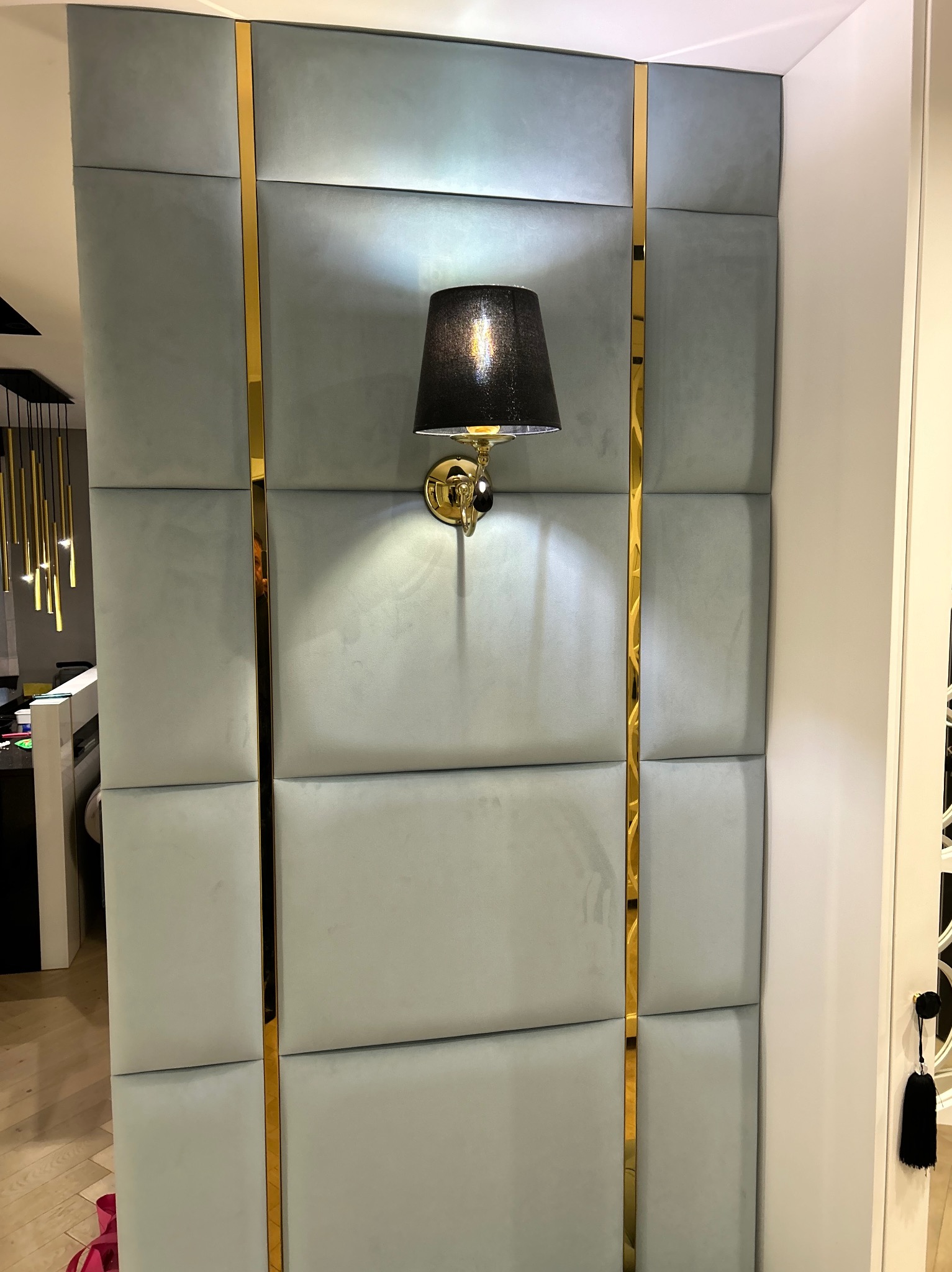 Panel ścienny z miękkiej, jasnoniebieskiej tapicerki, podzielony złotymi listwami, oświetlony kinkietem z czarnym abażurem