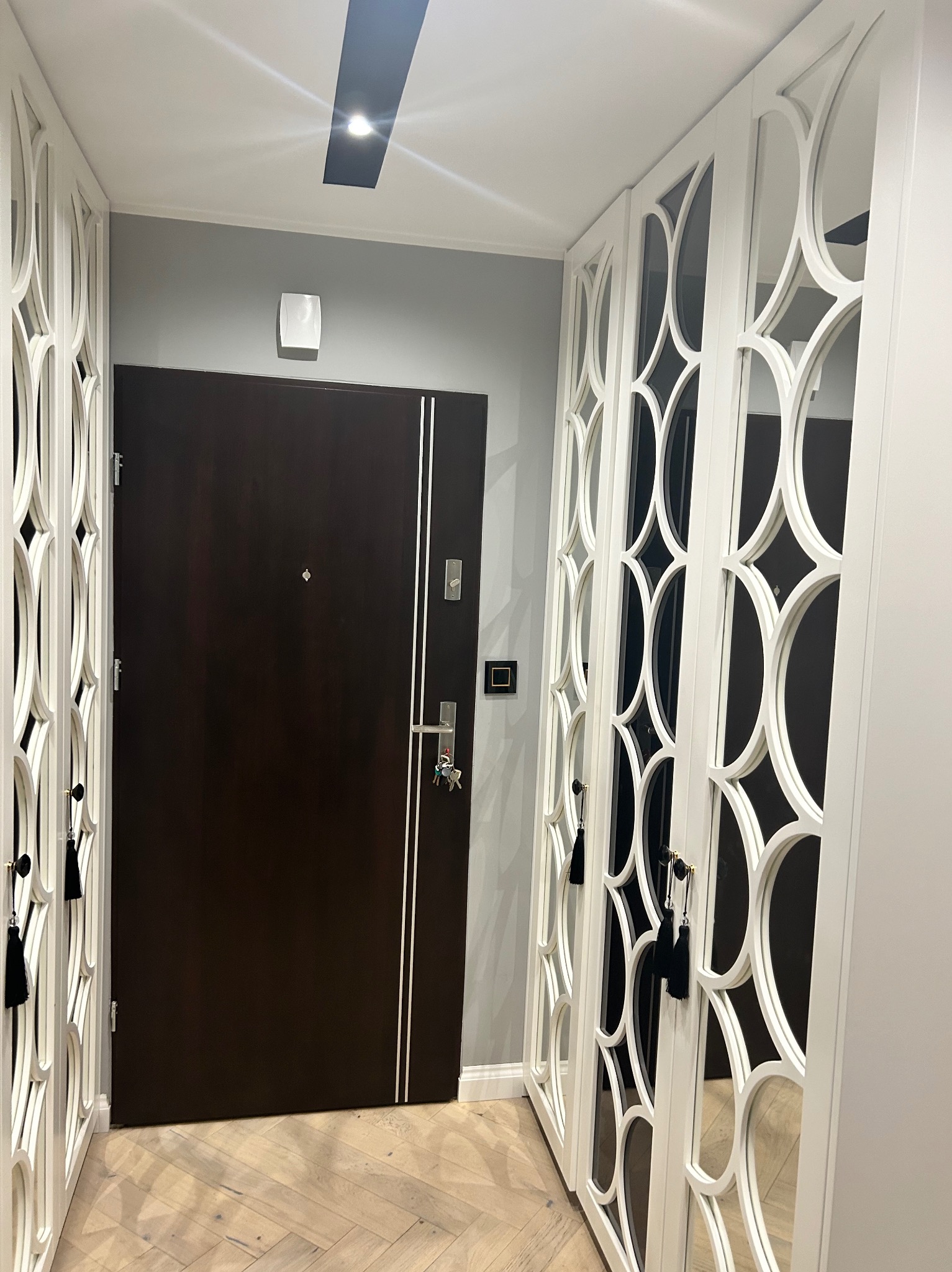 Przedpokój z zabudową szaf wnękowych z białymi, dekoracyjnymi frontami z motywem okręgów i ciemnymi lustrami, oraz brązowymi drzwiami wejściowymi z srebrnymi akcentami.