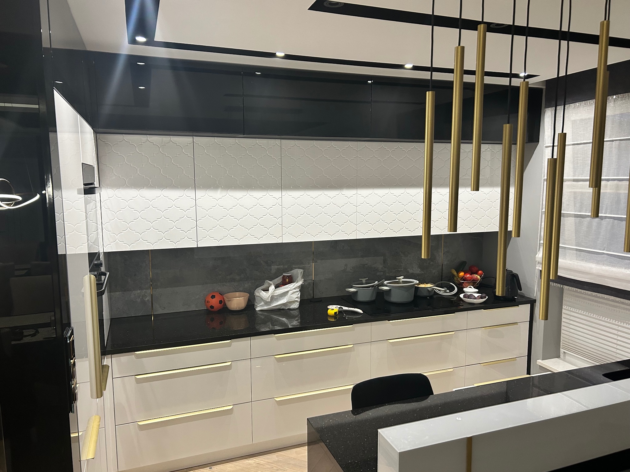 Nowoczesna kuchnia z białymi szafkami dolnymi z delikatnymi złotymi uchwytami, czarnymi szafkami górnymi i czarnym blatem, oświetlona złotymi lampami wiszącymi, widoczne naczynia i owoce na blacie.