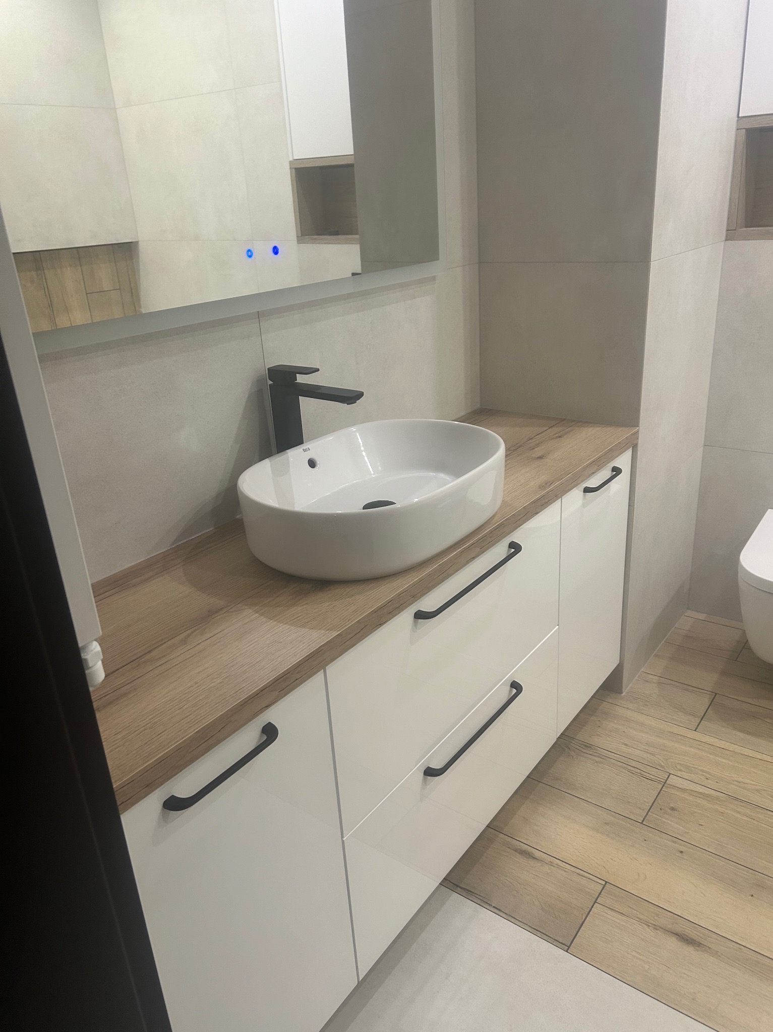 Nowoczesna łazienka: biała, owalna umywalka nablatowa z czarną armaturą, szafka pod umywalkę z białymi, lakierowanymi frontami i drewnianym blatem, drewnopodobne płytki na podłodze, duże lustro...