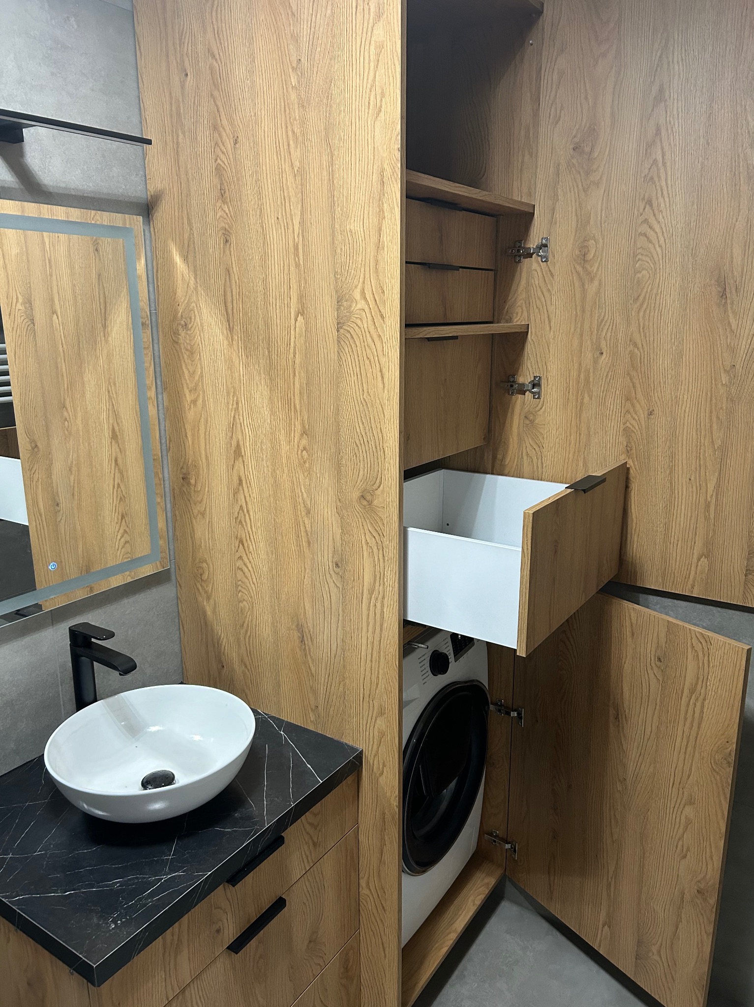 Szafa wnękowa z drewnianymi frontami kryjąca pralkę, obok umywalka nablatowa na czarnym blacie z marmurkowym wzorem, lustro z podświetleniem LED.