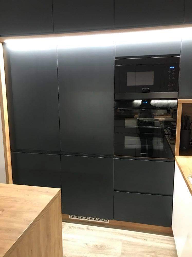 Nowoczesna zabudowa kuchenna w kolorze grafitowym z wbudowanym piekarnikiem i mikrofalówką Samsung, oświetlenie LED podszafkowe, drewniany blat wyspy w tle.