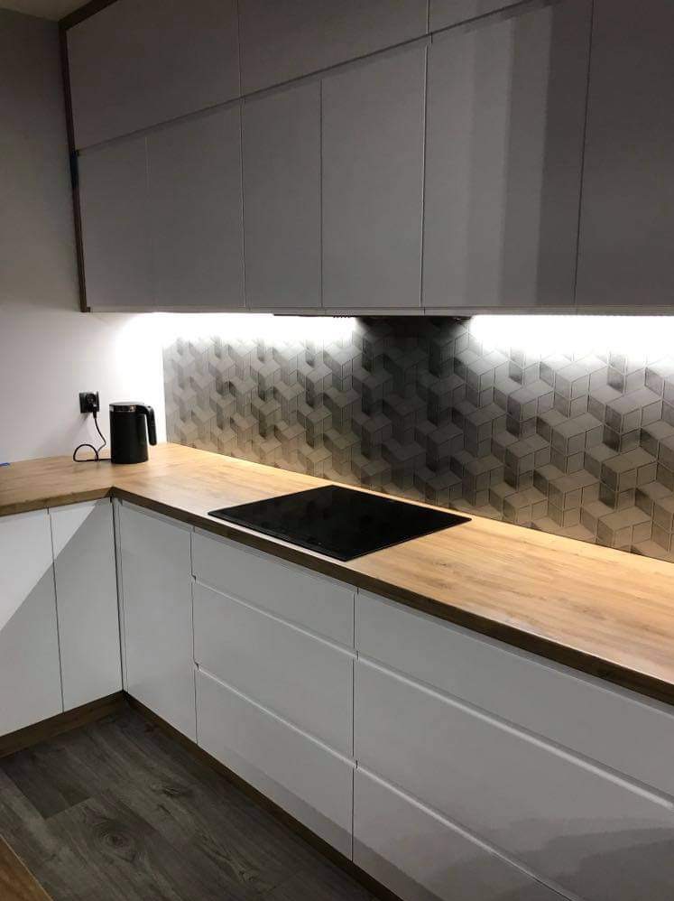 Nowoczesna kuchnia z białymi frontami szafek i drewnianym blatem, podświetlana listwą LED, na ścianie dekoracyjna mozaika z geometrycznym wzorem 3D, czarny czajnik elektryczny w rogu.