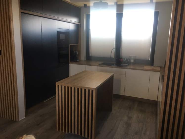 Nowoczesna kuchnia z czarnymi szafkami górnymi, białymi dolnymi i wyspą z drewnianych listewek. Widoczne okno z roletami i drewniane panele ścienne.