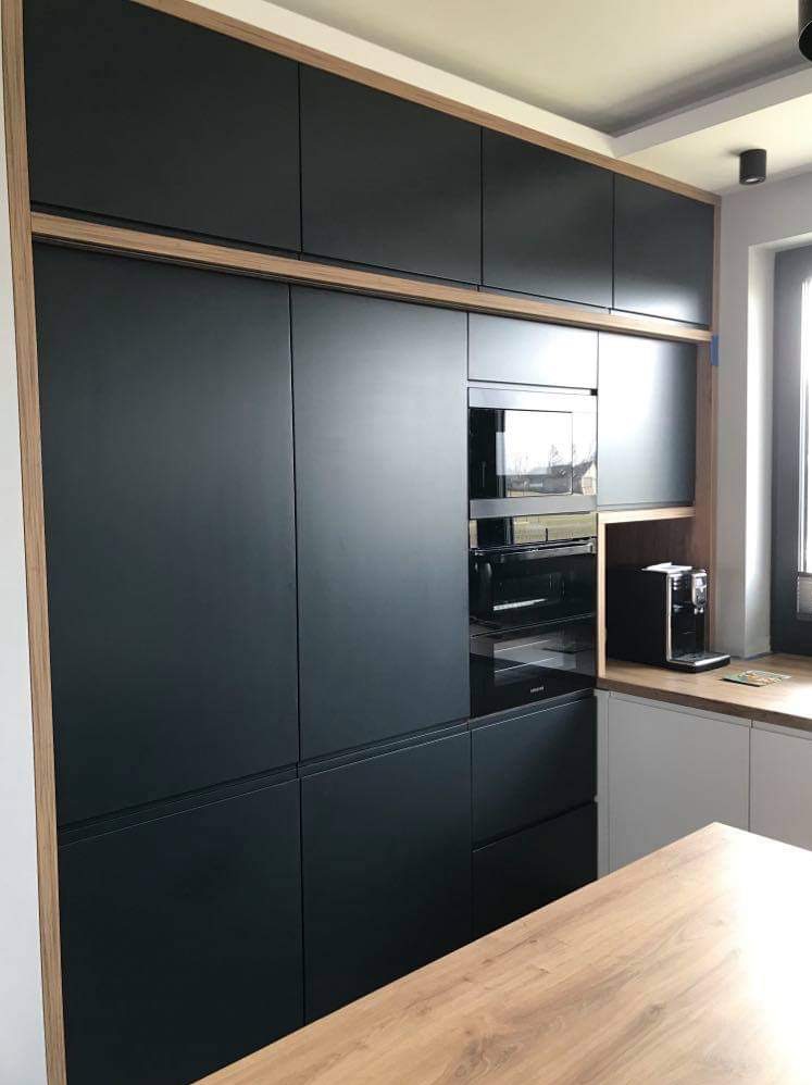 Czarna zabudowa kuchenna z drewnianymi akcentami, piekarnikiem i mikrofalówką w zabudowie, ekspres do kawy na blacie, minimalistyczny design.
