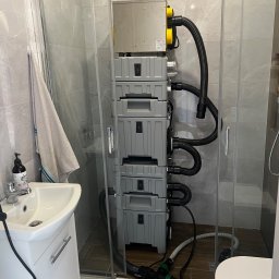 Sucha Strefa - Profesjonalny sprzęt do osuszania w kabinie prysznicowej z szarymi płytkami. Widoczne węże, armatura i elementy instalacji. Domowe warunki.