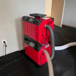 Czerwony, dwupoziomowy osuszacz kondensacyjny z wężami na czarnej macie. Urządzenie podłączone do gniazdka. Widoczne panele sterowania i etykieta producenta.