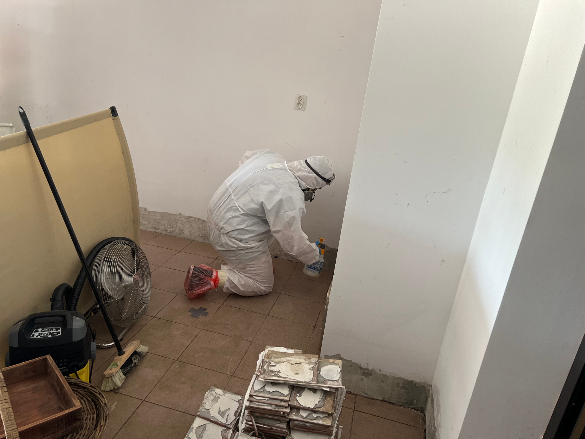 Pracownik w kombinezonie ochronnym klęczy i spryskuje ścianę preparatem, na podłodze leżą stare płytki i stoi wentylator
