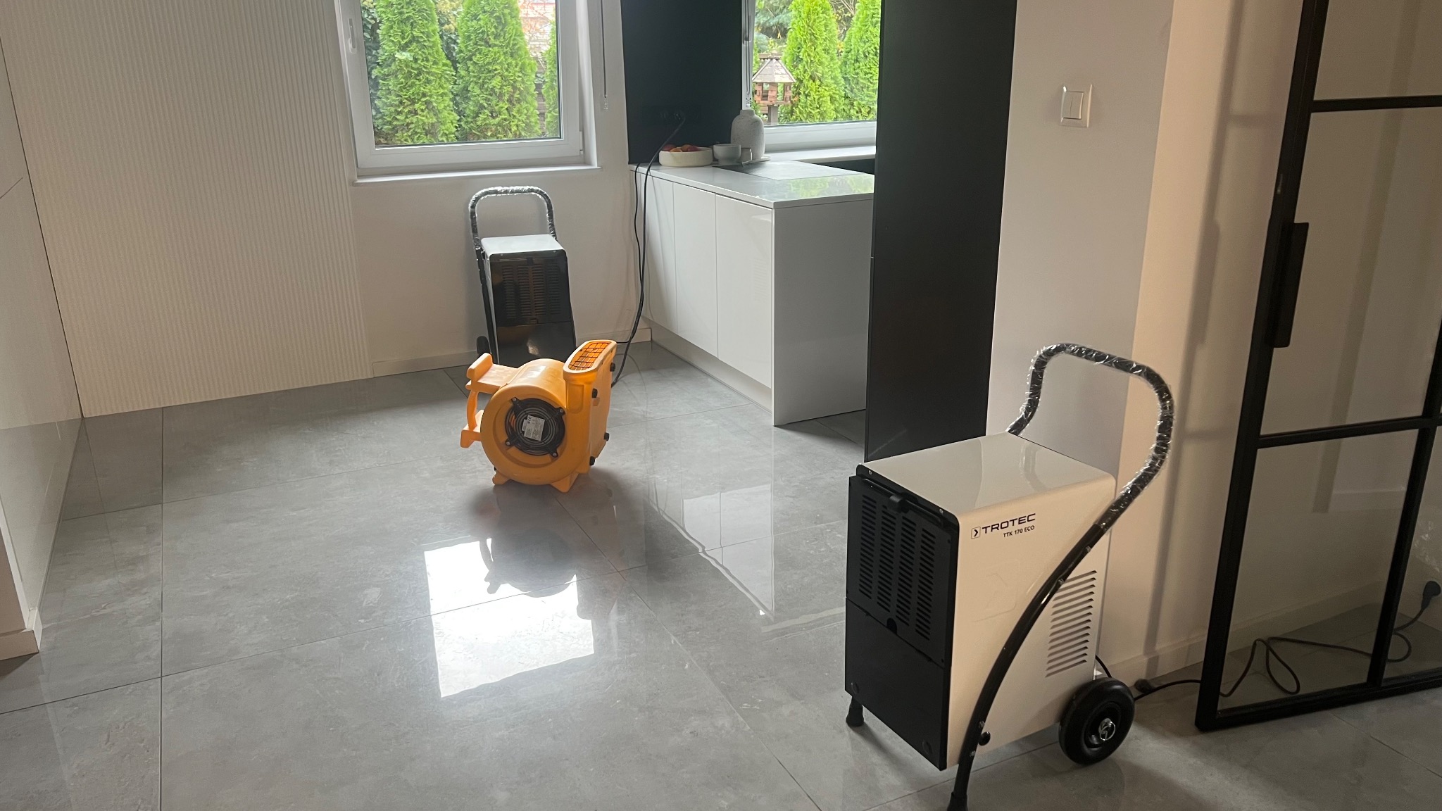 Dwa urządzenia do osuszania pomieszczeń na szarej podłodze: biały osuszacz Trotec TTK 170 ECO z czarnymi elementami i żółta dmuchawa powietrza w nowym pomieszczeniu z oknem.