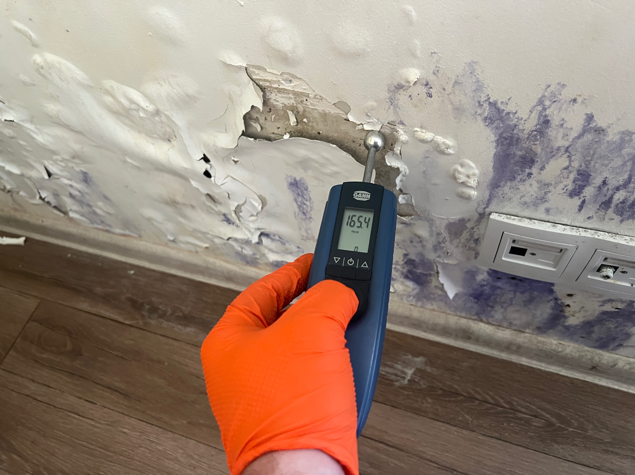 Pomiar wilgotności ściany z widocznymi śladami pleśni i odchodzącej farby, wykonany miernikiem w pomarańczowej rękawicy ochronnej.