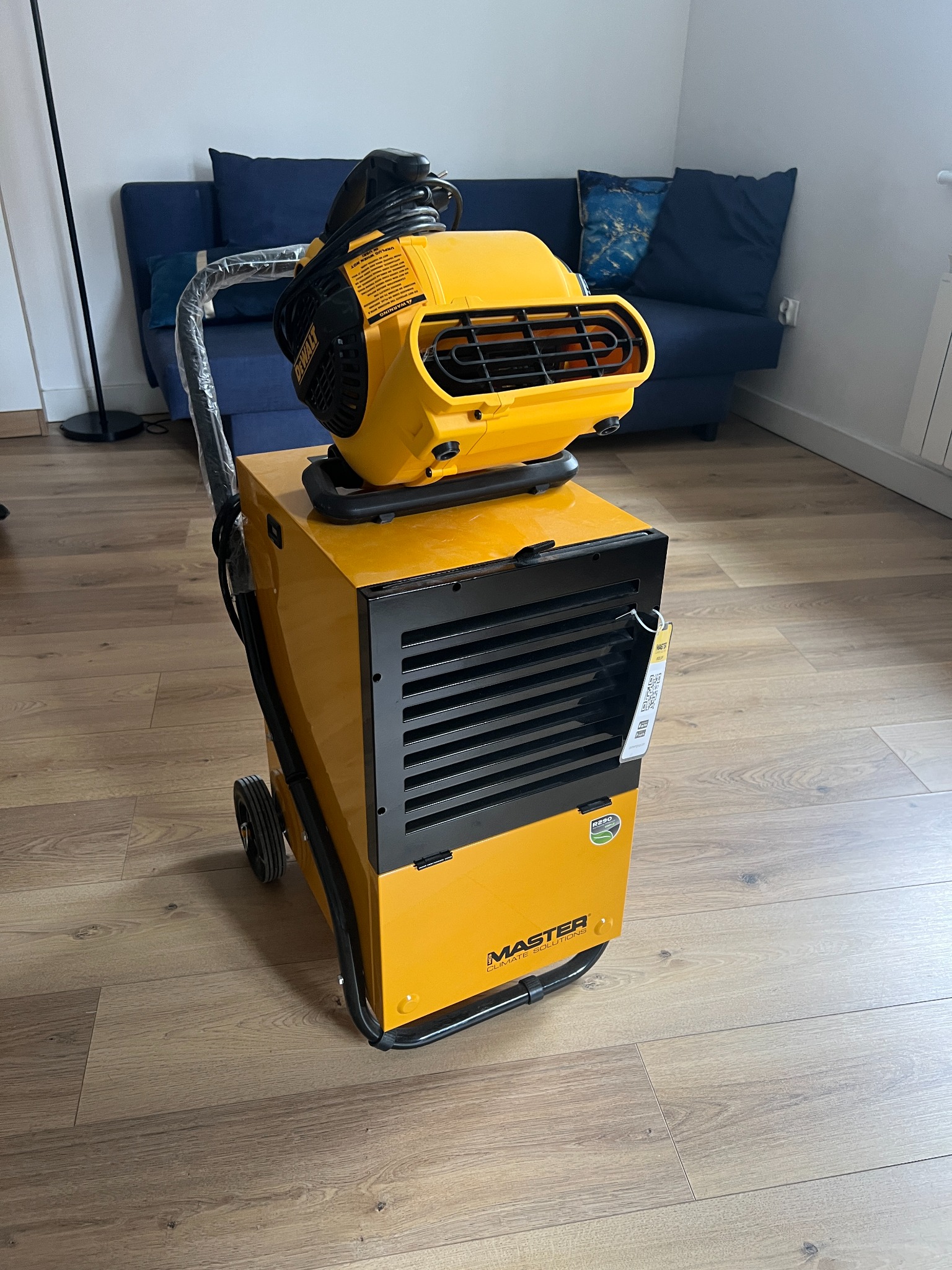 Żółty, mobilny osuszacz powietrza marki Master z czarnymi elementami na drewnianej podłodze, w tle niebieska sofa i biała ściana. Widoczne detale urządzenia.