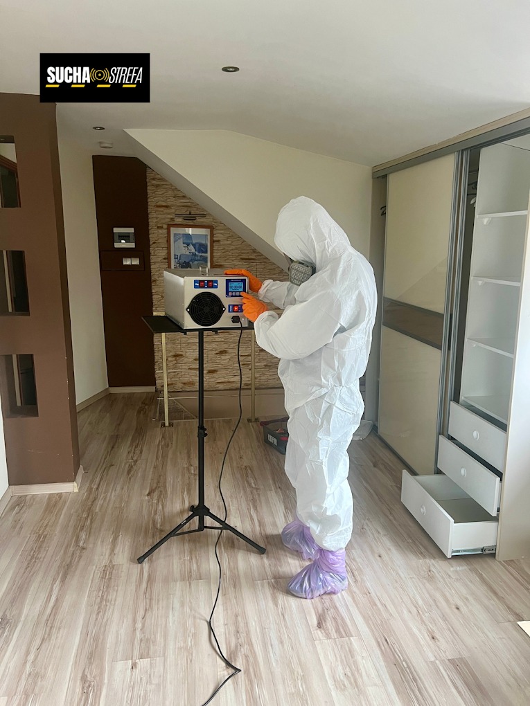 Pracownik w kombinezonie ochronnym obsługuje osuszacz powietrza na trójnogu w jasnym wnętrzu z drewnianą podłogą i szafą przesuwną. Widoczny logotyp firmy.