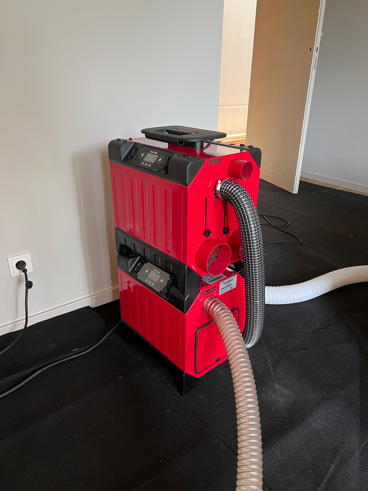 Czerwony, dwupoziomowy osuszacz kondensacyjny z wężami na czarnej macie. Urządzenie podłączone do gniazdka. Widoczne panele sterowania i etykieta producenta.