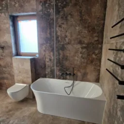 Nowoczesna łazienka z wolnostojącą wanną, umywalką i płytkami imitującymi kamień. Czarny grzejnik drabinkowy i minimalistyczna armatura.