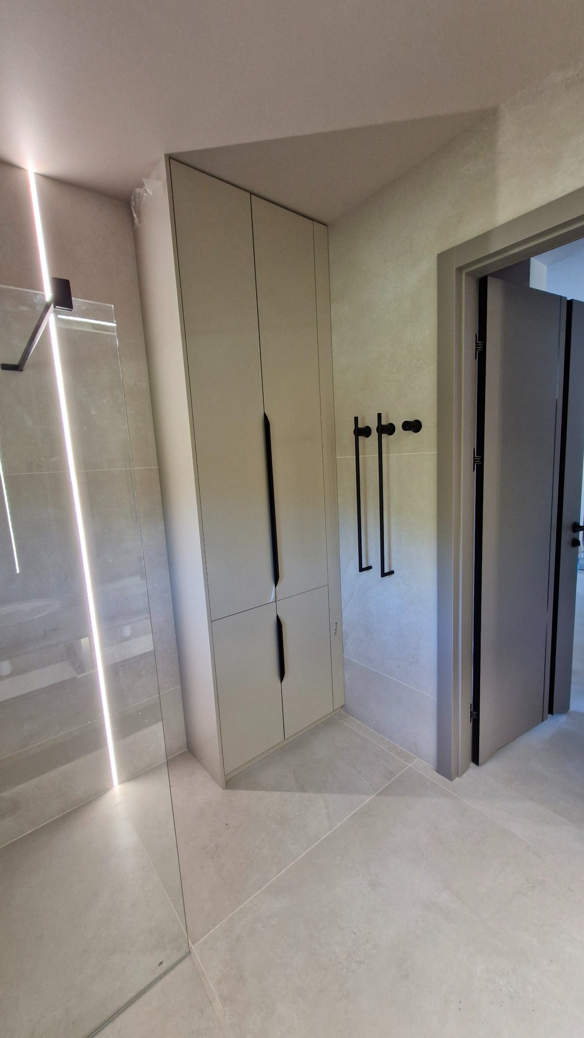 Nowoczesna łazienka z kabiną prysznicową typu walk-in, szafką z długimi uchwytami i szarymi płytkami podłogowymi oraz ściennymi. Minimalistyczny design.