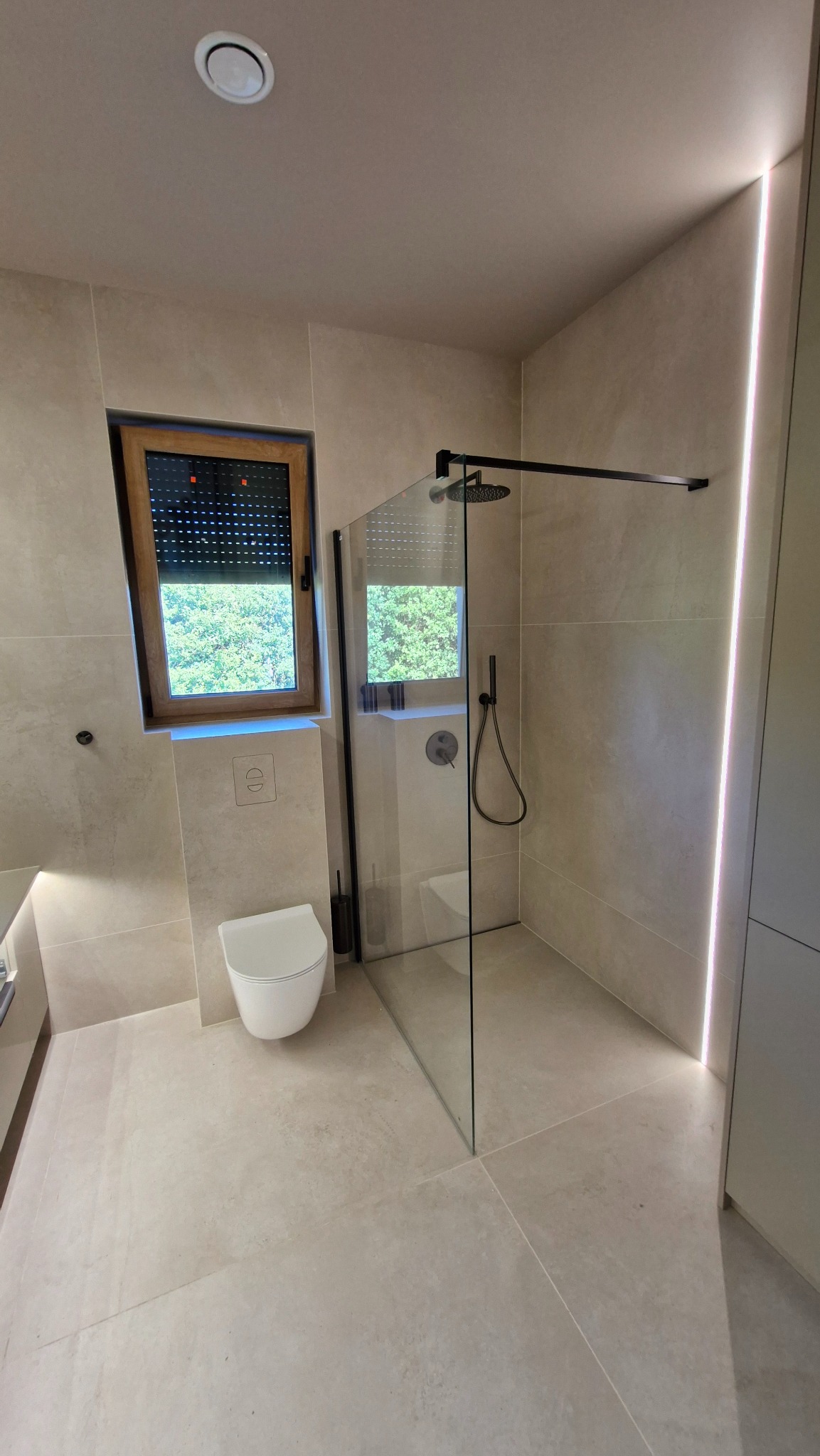 Jasna łazienka z kabiną typu walk-in, oknem z roletami i podświetleniem LED. Minimalistyczny design, beżowe płytki i czarne akcenty.