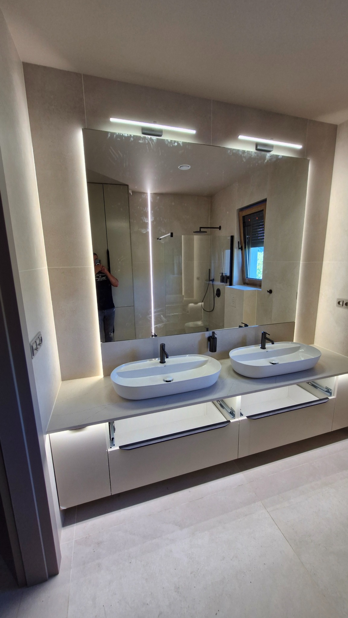 Nowoczesna łazienka z podwójną umywalką na blacie, podświetlana listwami LED, duże lustro z oświetleniem. Widoczne otwarte szuflady i beżowe płytki na ścianach.