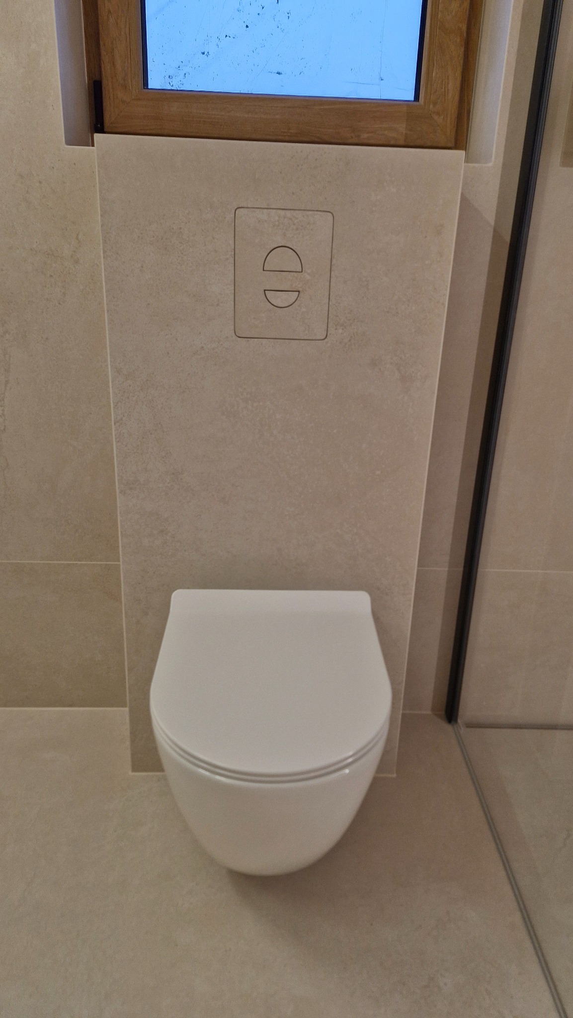 Podwieszana toaleta z beżowymi płytkami, w tle fragment okna i kabiny prysznicowej. Minimalistyczny design z przyciskiem spłukiwania w kształcie dwóch kół.