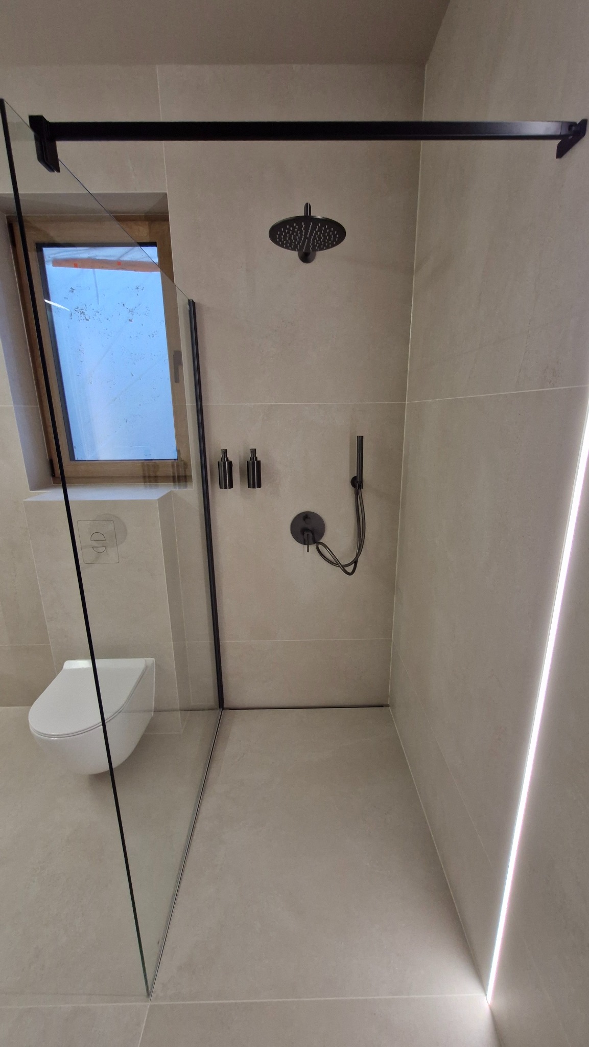 Nowoczesna łazienka z kabiną walk-in, czarną armaturą i podświetleniem LED. Minimalistyczny design, beżowe płytki. Widoczna toaleta pod oknem.