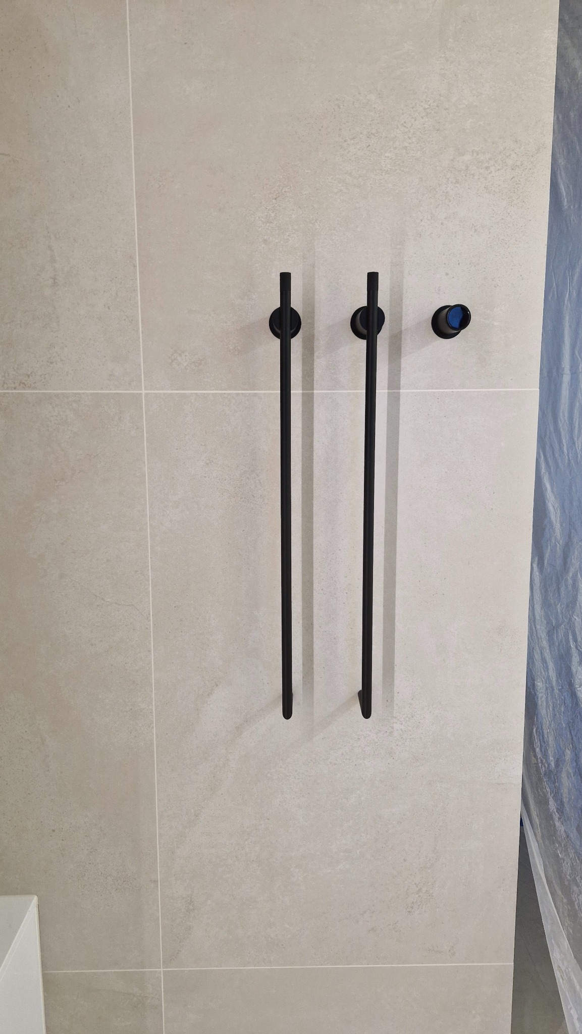 Beżowe płytki łazienkowe z czarnymi, minimalistycznymi uchwytami i elementem instalacji wodnej. Częściowo widoczne zabezpieczenie folią malarską.