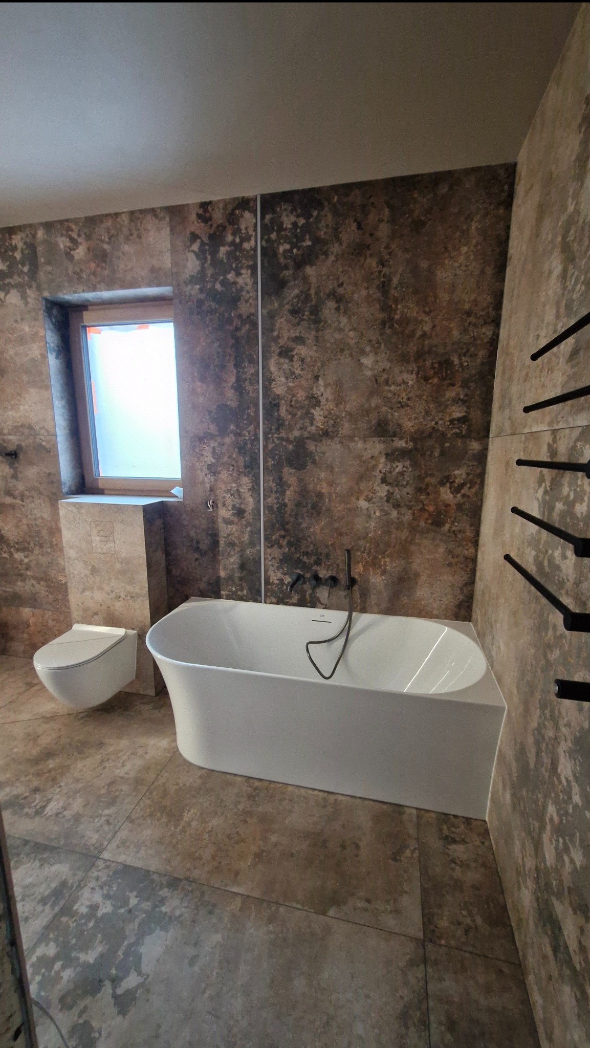 Nowoczesna łazienka z wolnostojącą wanną, umywalką i płytkami imitującymi kamień. Czarny grzejnik drabinkowy i minimalistyczna armatura.