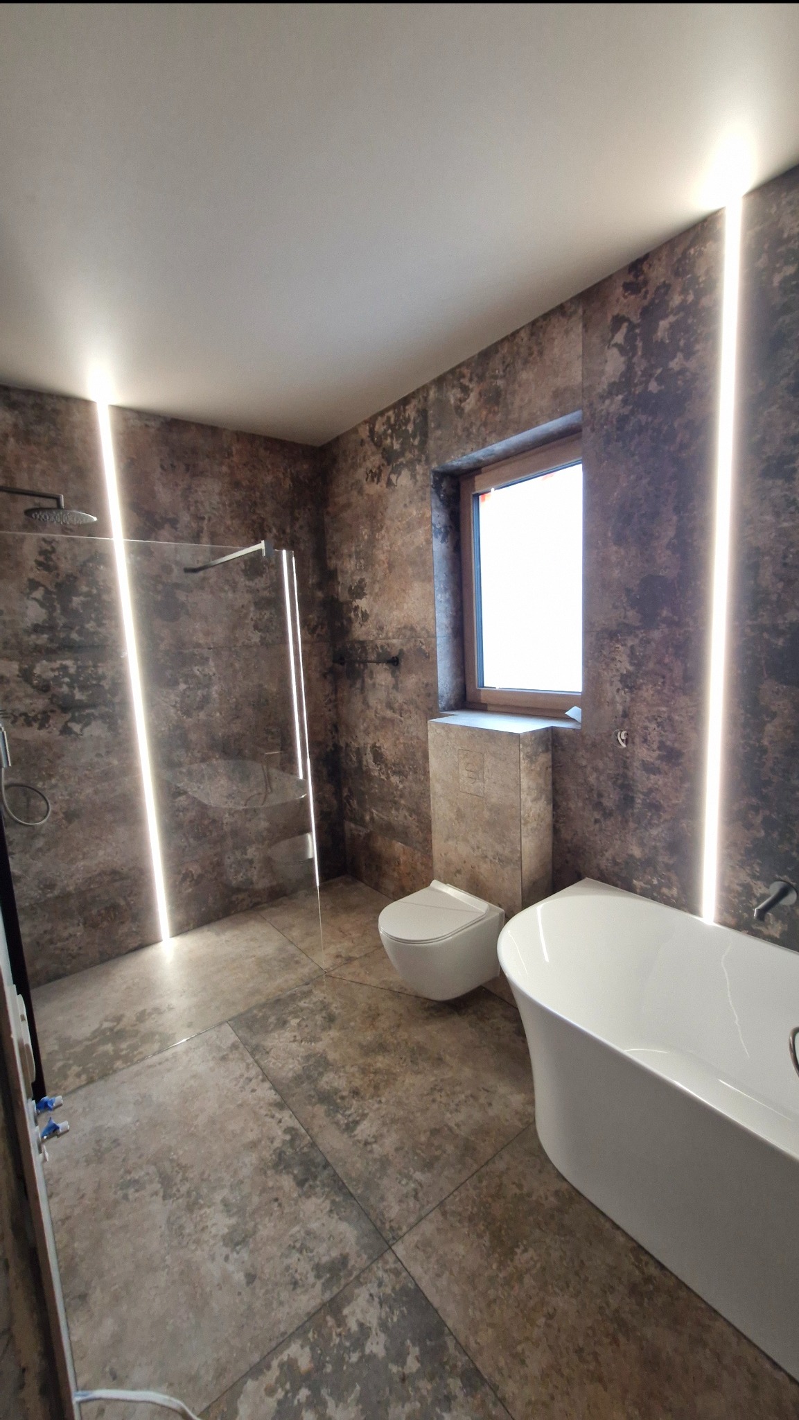 Nowoczesna łazienka po remoncie w Świdnicy. Kabina prysznicowa typu walk-in, wolnostojąca wanna i podwieszana toaleta. Płytki imitujące beton i pionowe pasy LED.