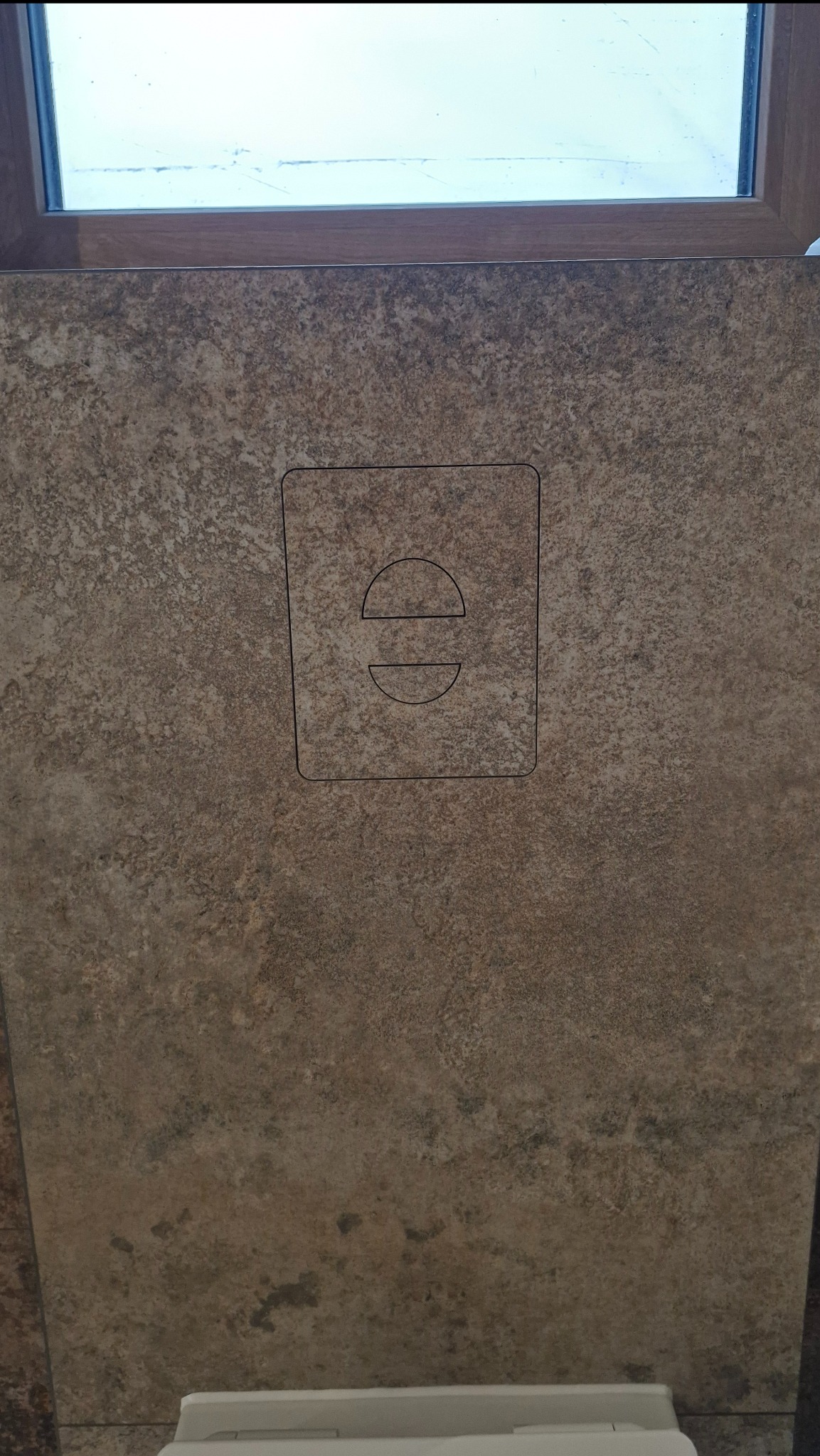 Beżowa płytka z wyciętym kształtem przycisku spłuczki, zamontowana w łazience pod oknem, widoczny fragment toalety. Wykończenie łazienki w neutralnej tonacji.