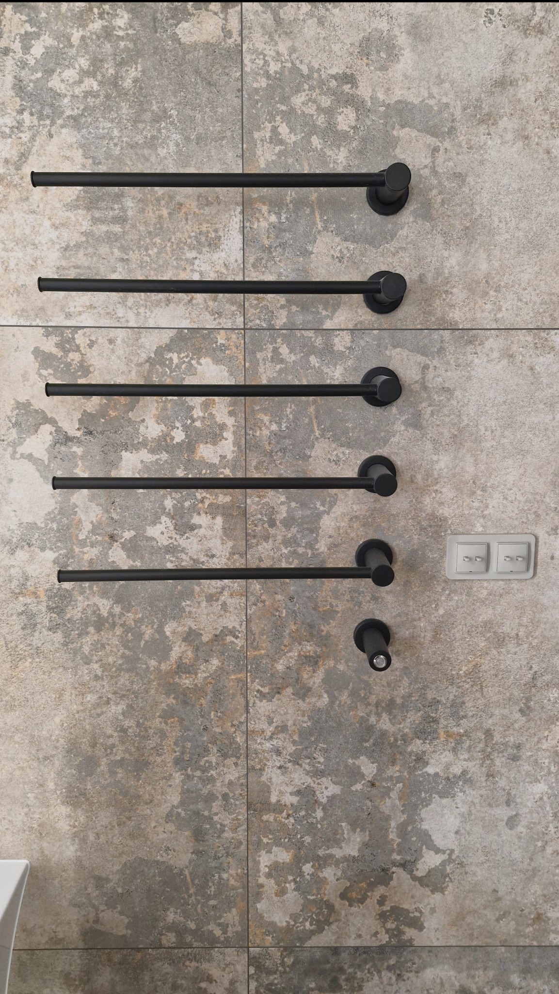 Ściana z płytek imitujących kamień z zamontowanym czarnym, drabinkowym grzejnikiem łazienkowym i włącznikiem światła. Minimalistyczny design.