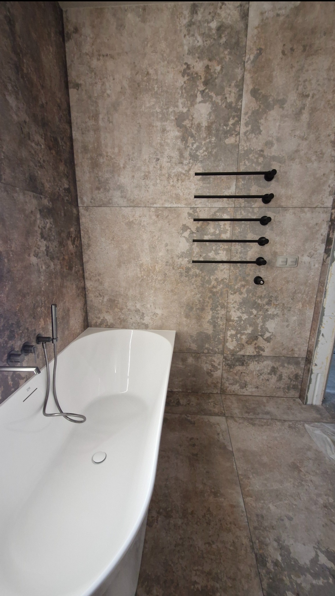 Nowoczesna łazienka z wolnostojącą wanną i ścianami wyłożonymi płytkami imitującymi beton. Czarny grzejnik drabinkowy dodaje elegancji.