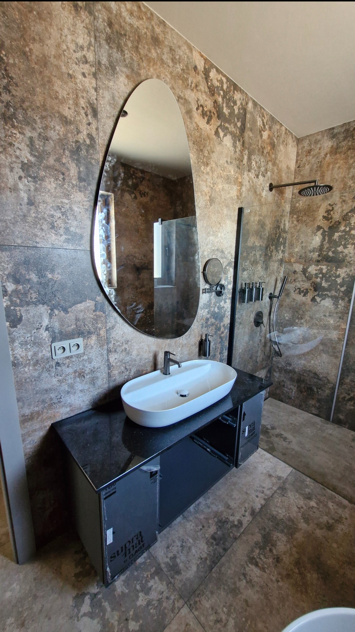 Nowoczesna łazienka z owalnym lustrem i umywalką nablatową. Ściany i podłoga wykończone płytkami imitującymi kamień. Widoczna kabina prysznicowa ze szklaną ścianką.